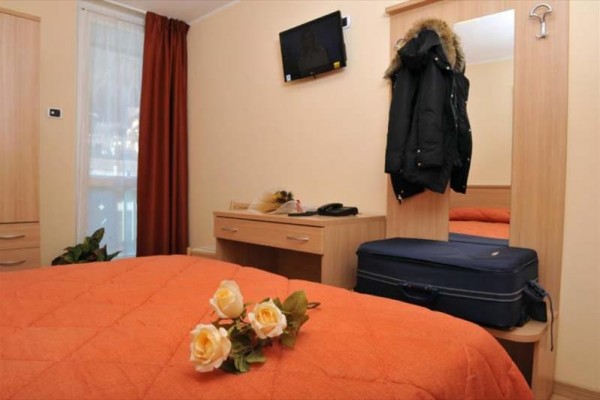 Pokój hotelowy z pomarańczową narzutą na łóżku, różami i bagażem przy lustrze.