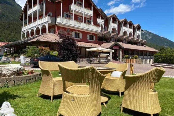 Hotel z czerwoną fasadą i rattanowymi meblami na trawniku na pierwszym planie, otoczony górami.