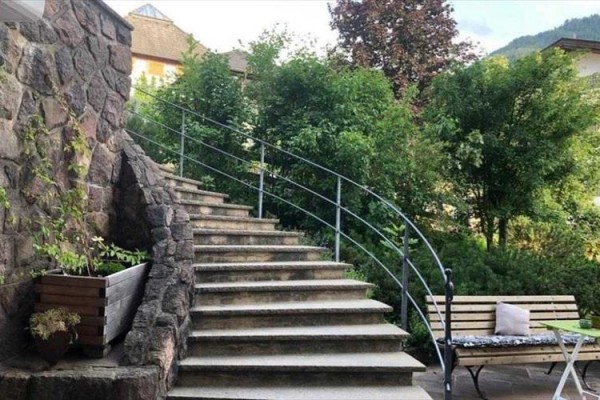 Kręte kamienne schody prowadzące w górę między zielonymi krzewami, obok ławka i stolik.